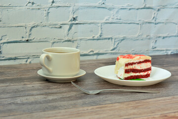A cup of hot black coffee on a saucer, a fork and a plate with a piece of red velvet cake on a wooden table.