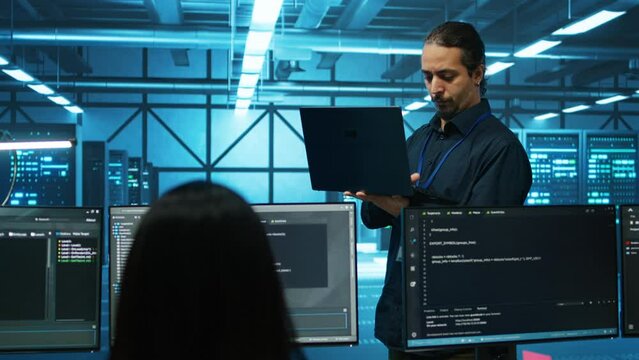 Engineer typing on laptop in server hub, checking recovery plan, monitoring data center energy consumption across components. Technician inspecting rackmounts, doing routine disk checks