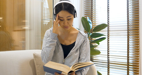 stress asian woman use headphones listen to music reading book sitting on cozy sofa in living room...