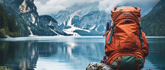 Traveler with an orange backpack sitting by a serene mountain lake, surrounded by lush green forests and picturesque snowy peaks. - Powered by Adobe