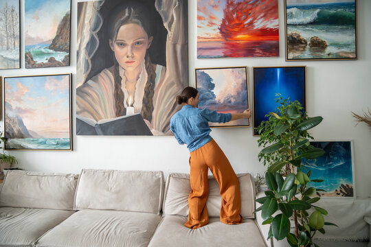 Interested woman painter, hangs on wall trendy artwork, created handmade in art workshop. Inspired pensive female artist designer holds new finished painting ponder compose for decoration art studio.