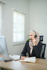 A woman wearing a hijab headset is talking on the phone while sitting at a desk