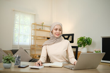 A woman wearing a head scarf sits at a desk with a laptop and a notebook