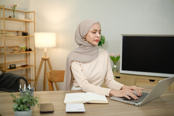 A woman wearing a scarf sits at a desk with a laptop and a notebook