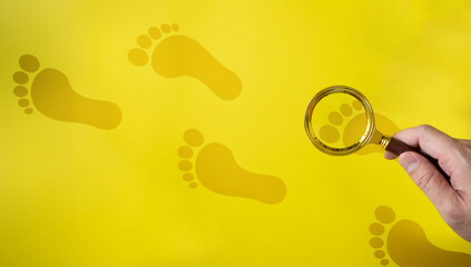 Human hand holding a magnifying glass looking at human footprints on a yellow background. Concept...