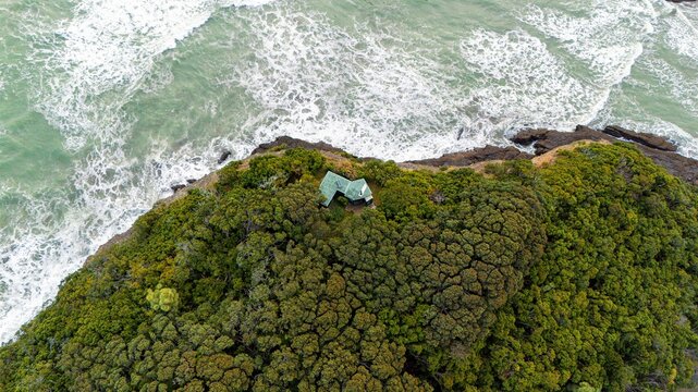 An aerial shot of a secluded cabin nestled in a dense coastal forest, surrounded by the ocean waves and rocky shore  - Bethells Beach