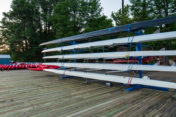A row of rowing boats is neatly arranged on racks along a wooden boat dock, while kayaks are...