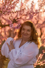 Woman blooming peach orchard. Against the backdrop of a picturesque peach orchard, a woman in a long white dress enjoys a peaceful walk in the park, surrounded by the beauty of nature.