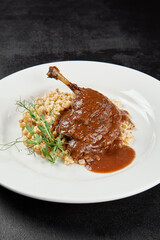 Classic dish french cuisine - roasted duck leg in porto sauce with garnish on black background. Cooked duck leg with pearl barley on white plate on dark concrete table. Duck confit in minimal style.