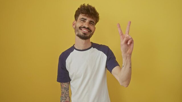 Young, happy hispanic man confidently standing over an isolated yellow background, smiling and pointing up with two fingers to signify the number two