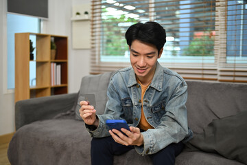 Smiling young Asian man doing online payment through credit card and smart phone