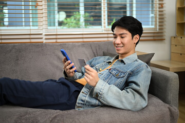 Pleased young adult man holding credit card and using mobile application for online shopping