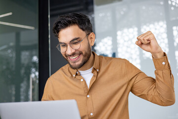 Fototapeta premium Happy man celebrating success while working on laptop in modern office