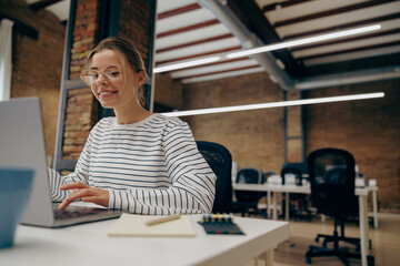 Smiling young woman manager working on laptop while sitting the desk in stylish office