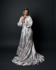 portrait of beautiful blonde female model wearing romantic historical gown white bridal floral pattern. standing pose  with flowing silk skirt. isolated dark studio background.