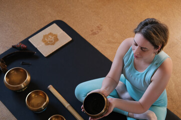 A yogi girl plays on a Tibetan singing bowl. A young woman meditating in yoga on the floor in the...