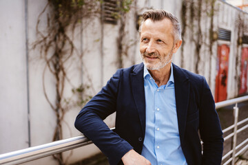 A confident mature businessman in a blue suit and shirt relaxes outdoors by an urban building's railing.