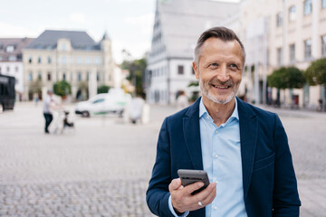 Happy senior businessman holding a smartphone while standing in a city square. Urban setting with...