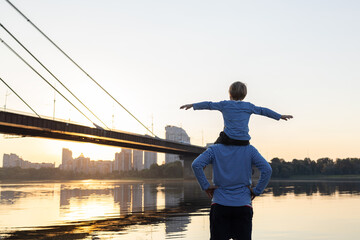 dad and son in identical long sleeves watching the sunset by the river. the child sits on the man s shoulders with his arms out to the side. Father's day, son's day. Tender relationships in the family