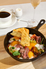 Sunny-side-up eggs in a skillet with roast beef, vegetables, and ciabatta, Americano coffee, and a glass of sparkling wine