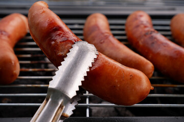 Metal tongues holding grilled sausage in front of bbq grill with many sausages.