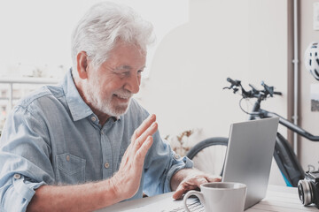 Video call concept. Portrait of smiling senior man sitting outdoor on a white table using laptop...