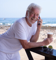 Cheerful bearded senior retired man outdoors at the seaside beach talking on mobile phone enjoying free time and relaxed vacation looking at camera. Horizon over water