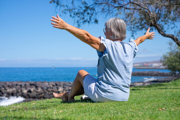 Back view of senior woman sitting barefoot in meadow fae the sea looking at the horizon with open arms, relaxed senior lady enjoying free time summer vacation or retirement
