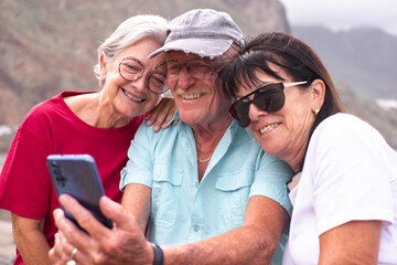 Group of cheerful senior Caucasian friends in leisure travel at sea sitting looking together at mobile phone. Two women and one man smiling enjoying retirement and free time in vacation
