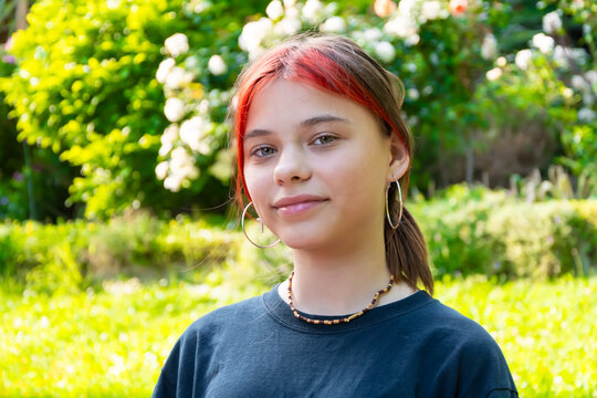 Portrait of a 12-year-old teenage girl with red hair and hoop earrings against a backdrop of a lush, green garden with blooming flowers on a sunny day.