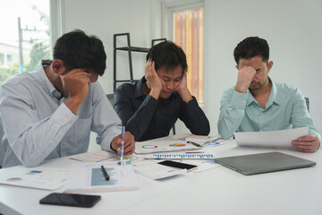 A team of tired, stressed young businessmen hold their heads in their hands. From working with problems without success Financial documents in the Ahimsarimsap project Office syndrome concept.