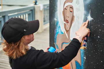 Female artist in black cap is painting picture with paint spray can spraying it onto canvas at outdoor street exhibition, side view of female art maker