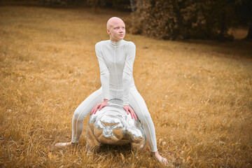 Young hairless girl with alopecia in white cloth sits on tardigrade figure in fall park, surreal scene with bald teenage girl confidently embraces her unique beauty