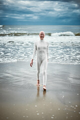 Smiling hairless girl with alopecia in white futuristic suit comes out of cold restless sea on sandy beach, metaphoric performance of bald female artist about overcoming challenges and self acceptance