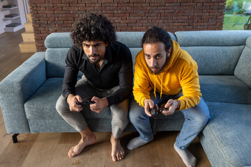 Two men are comfortably seated on a couch, engaging in a video game together, enjoying a leisure...