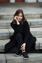 Attractive Caucasian young woman in a black coat sitting on the stair while walking along the city street on autumn day. Outdoors portrait. Travel, rest, fashion, style concept.