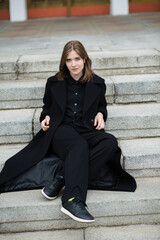 Attractive Caucasian young woman in a black coat sitting on the stair while walking along the city street on autumn day. Outdoors portrait. Travel, rest, fashion, style concept.