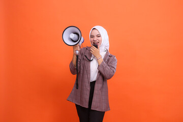 Angry young Asian woman in hijab facing right side to camera shouting using mic holding megaphone speaker isolated on orange background, Speech, announcing, advertising, broadcast concept