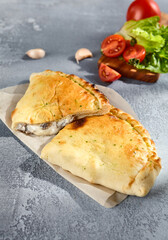 Golden calzone pizza on parchment paper, with fresh tomatoes and garlic in the background on a textured grey surface