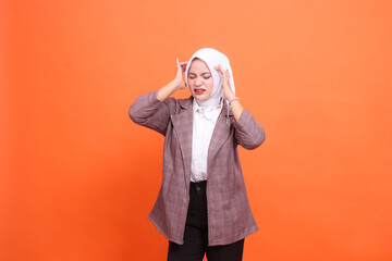 Beautiful young Asian girl wearing a hijab grimaces in pain, closes her eyes, experiences pain, dizziness, positions both hands on her head with an orange studio background. health care, advertising