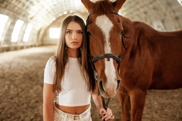 In white clothes, standing. Beautiful young woman is with horse indoors