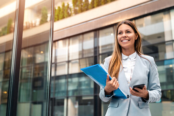 Young adult smiling professional business woman wearing suit holding smartphone and folder with...