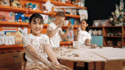 Cute girl smiling and sitting while diverse friend doing pottery workshop. Highschool student wearing dirty shirt while looking at camera at workshop in art lesson. Blurring background. Edification. - Powered by Adobe