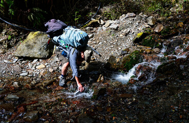 A tramper or hiker is leaning over refilling his stainless steel drinking bottle from a stream.