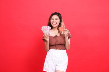 Asian girl laughing holding rupiah money and debit credit card wearing brown blouse top with red...