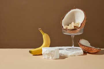 Frontal shot photograph against brown background, a few empty plaster platforms containing a glass with half a coconut and many banana slices placed on top, a fresh banana decoration next to it