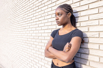 A pensive young woman with braids and crossed arms is leaning against a brick wall in an urban...