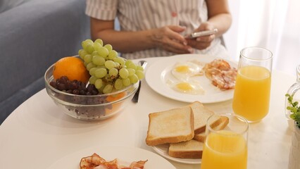 During breakfast, a woman uses her phone while enjoying her food. The breakfast table is set with scrambled eggs and juice while she checks her phone. Ideal for breakfast and technology topics.