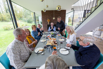 A group sharing a meal around a dining table in a modern home with large windows, creating a...