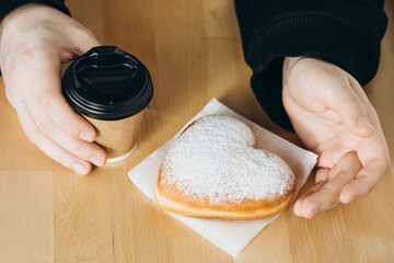 A paper cup of coffee and a heart-shaped donut in male hands, close-up.